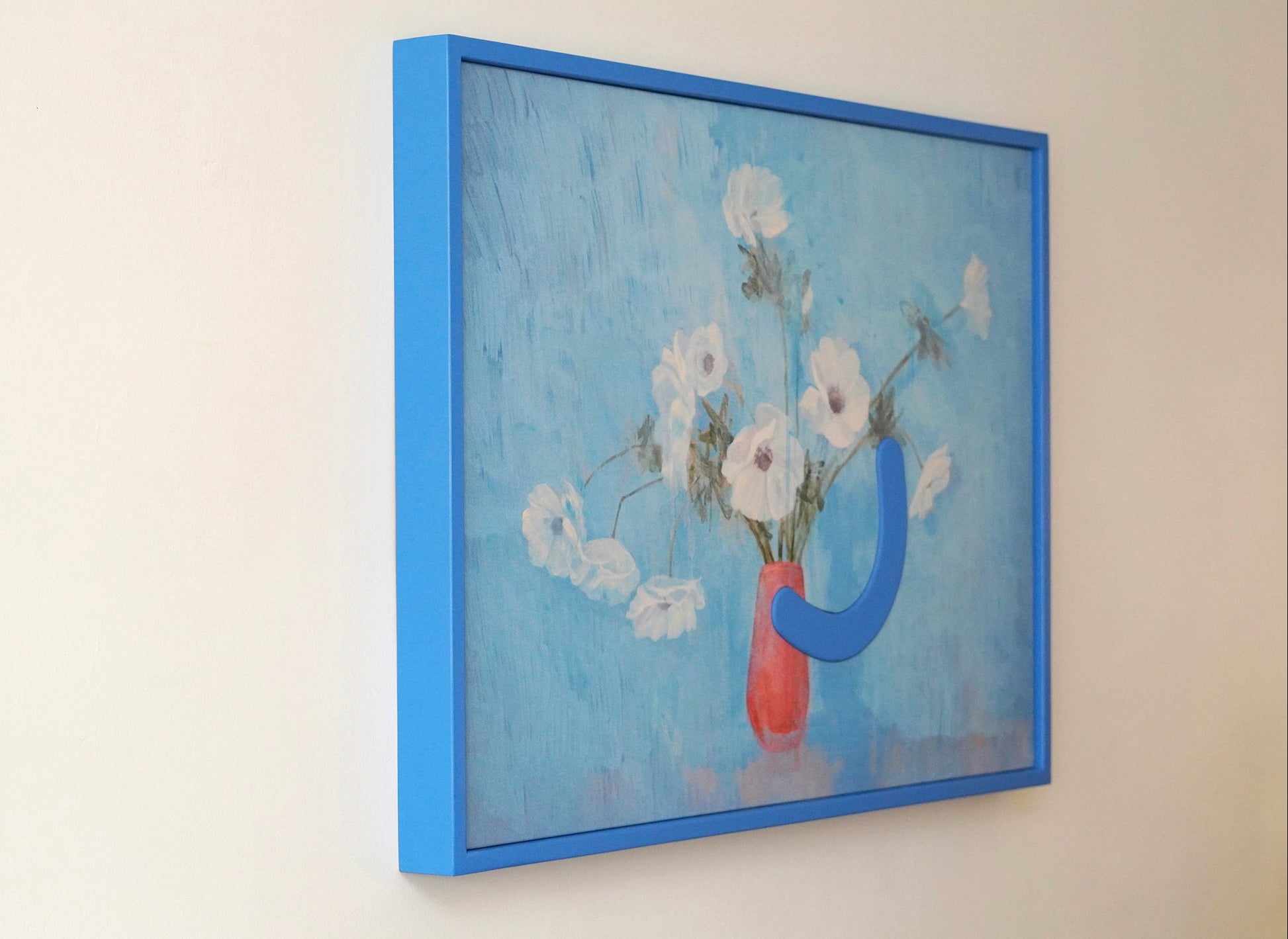 Framed artwork of a blue abstract sculpture holding a red vase with flowers on a light beige wall.